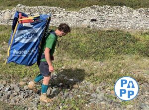 picture showing large 1 meter cubed bulk bag that has been converted in to a rucksack and is being used to carry plastic waste along a beach to a road head the beach is cobbled / pebbled with a dry stone wall shown to the rear of the beach